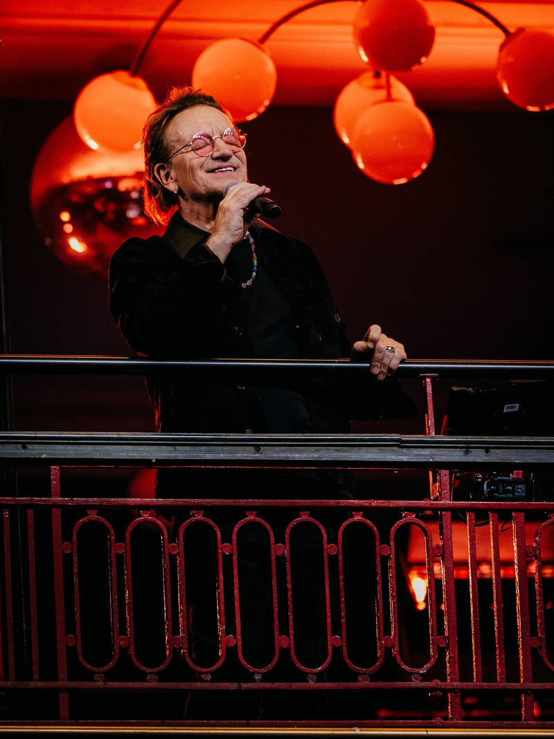 A man wearing round, reddish-orange tinted glasses and a black shirt stands on stage holding a microphone.