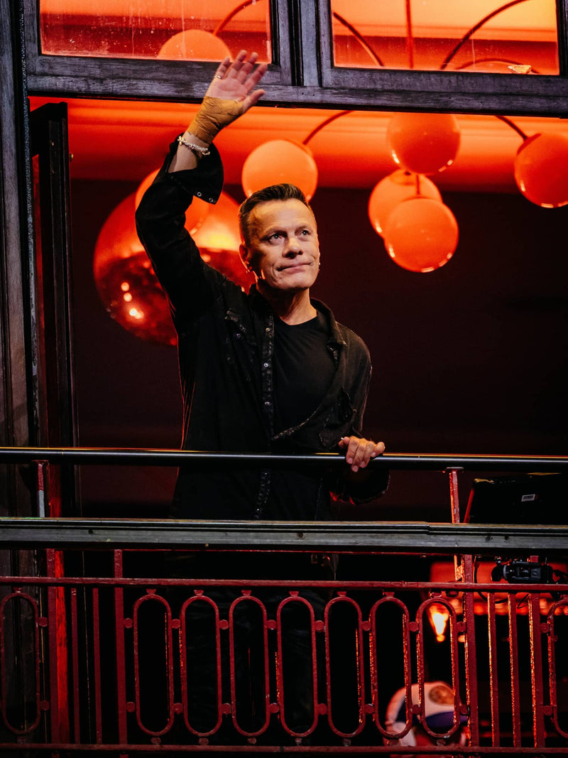 A man in a black jacket with intricate detailing waves from a balcony under warm orange lighting.