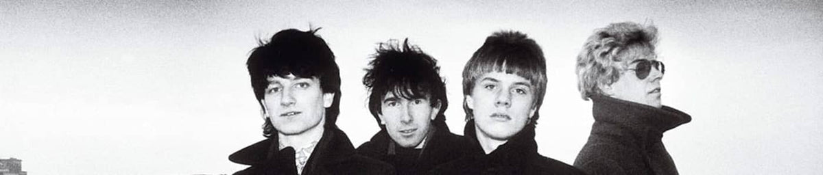 Black-and-white portrait of four men in dark coats, styled with 1960s fashion.